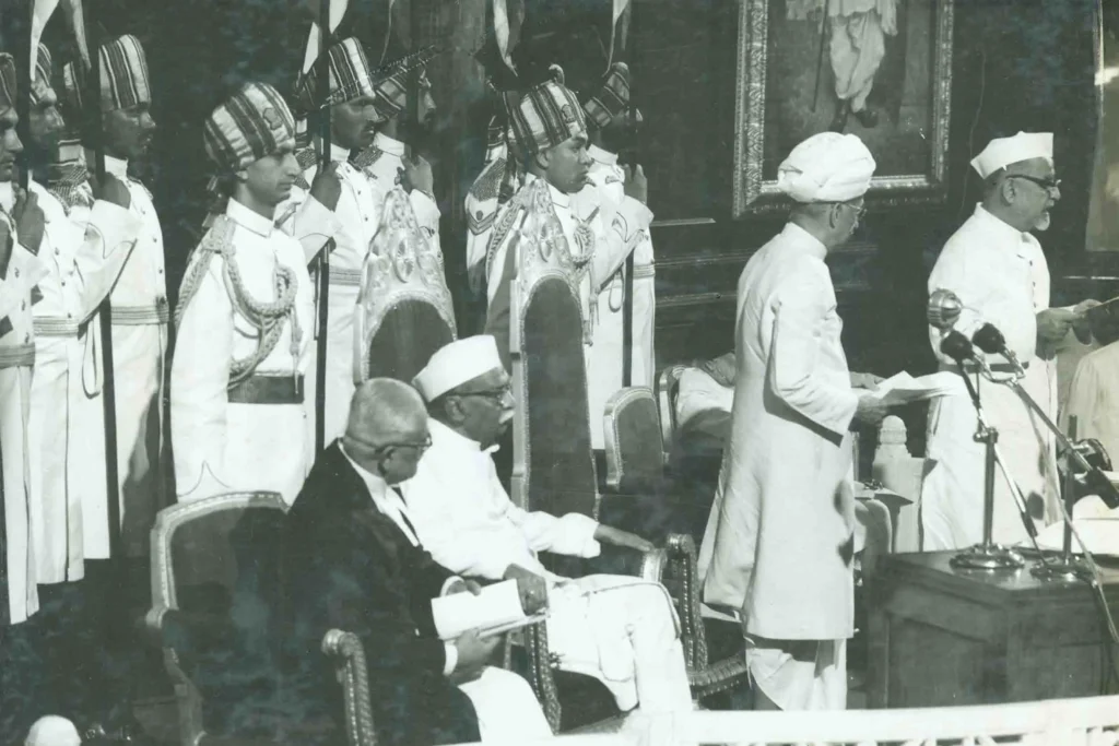 Dr. Sarvepalli Radhakrishnan standing and reading from a document during a formal swearing-in ceremony, with officials seated nearby and guards in ceremonial uniform standing behind.