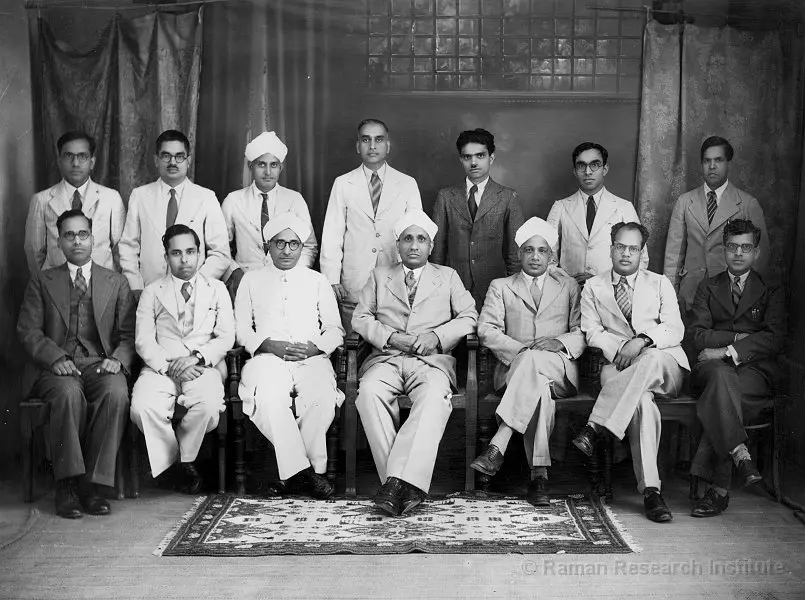 Group photograph of C. V. Raman with colleagues and researchers, all dressed in formal suits, seated and standing in rows inside a studio setting.