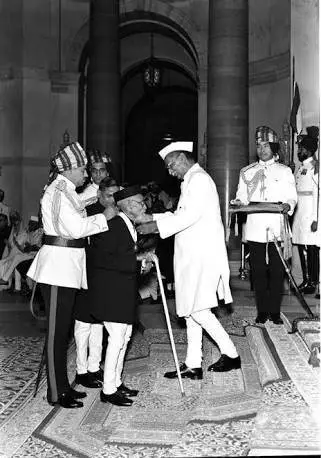 an elderly Maharshi Dhondo Keshav Karve, leaning on a cane, being honored with the Bharat Ratna by a dignitary (likely the President) in a formal ceremony; two uniformed attendants stand nearby in an ornate ceremonial hall.