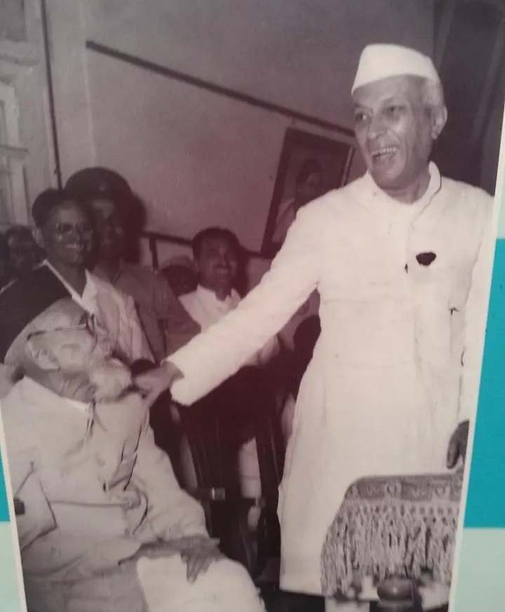 photograph showing Pandit Jawaharlal Nehru, standing and smiling in traditional attire (achkan and Gandhi cap), reaching out to touch the head of a seated Maharshi Dhondo Keshav Karve, with an audience visible in the background