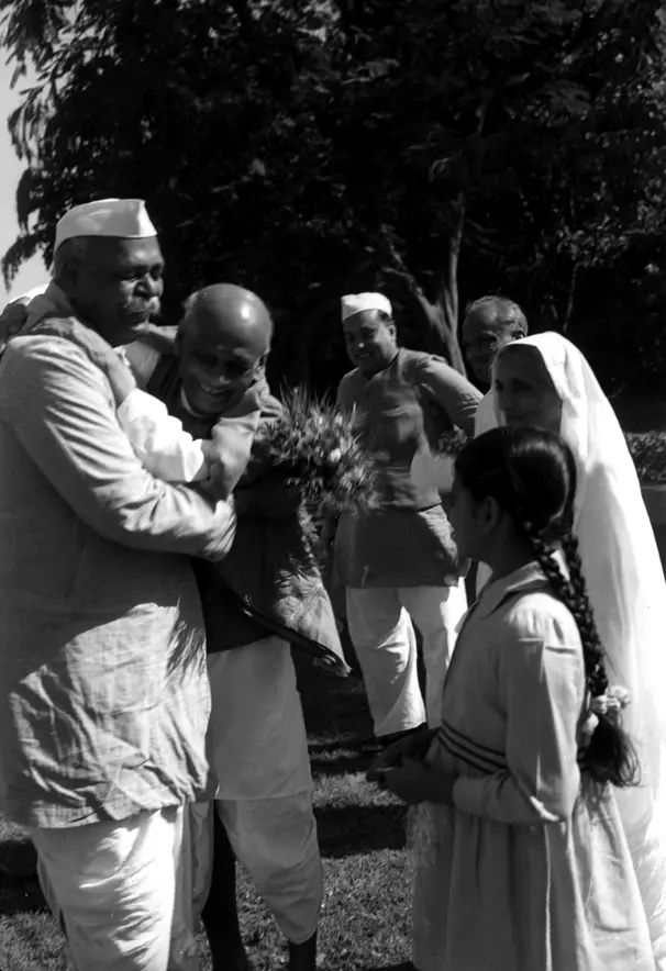 Govind Ballabh Pant warmly embracing Sardar Vallabhbhai Patel, with others including a woman and a young girl standing nearby, holding flowers in an outdoor setting.