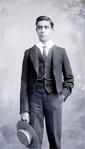 a young man in a three-piece suit holding a straw boater hat in one hand, with the other hand in his pocket, looking slightly to the side.