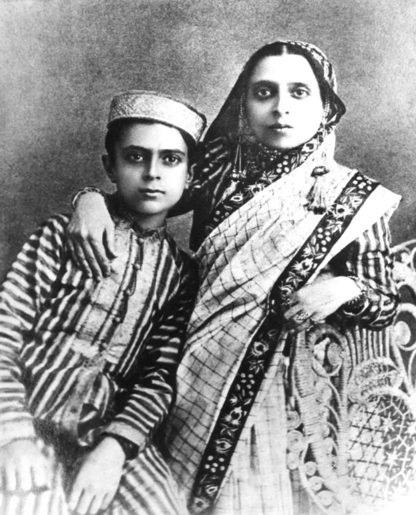 portrait of a young boy in traditional attire sitting beside a woman in a sari with an embroidered border, her arm around his shoulders, both looking directly at the camera.