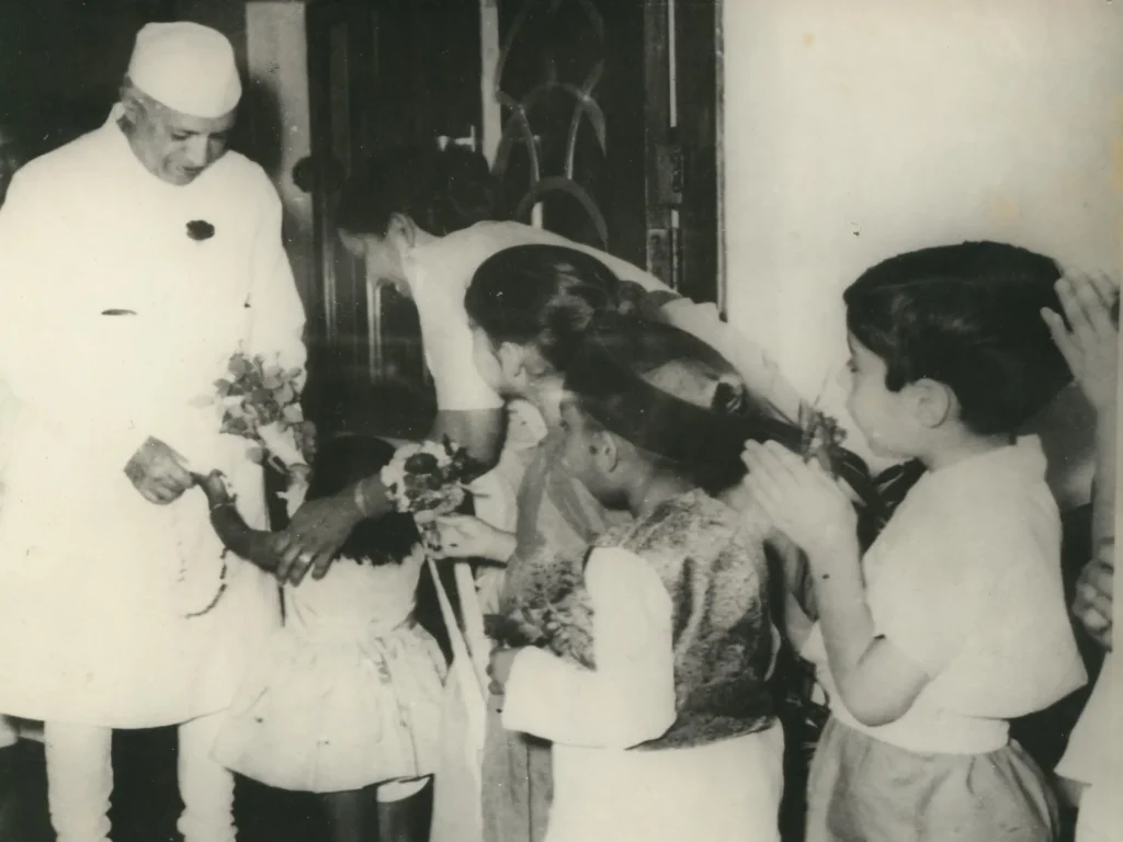The man is smiling and holding a bouquet while interacting warmly with the children, who are standing in a line and showing respect.
