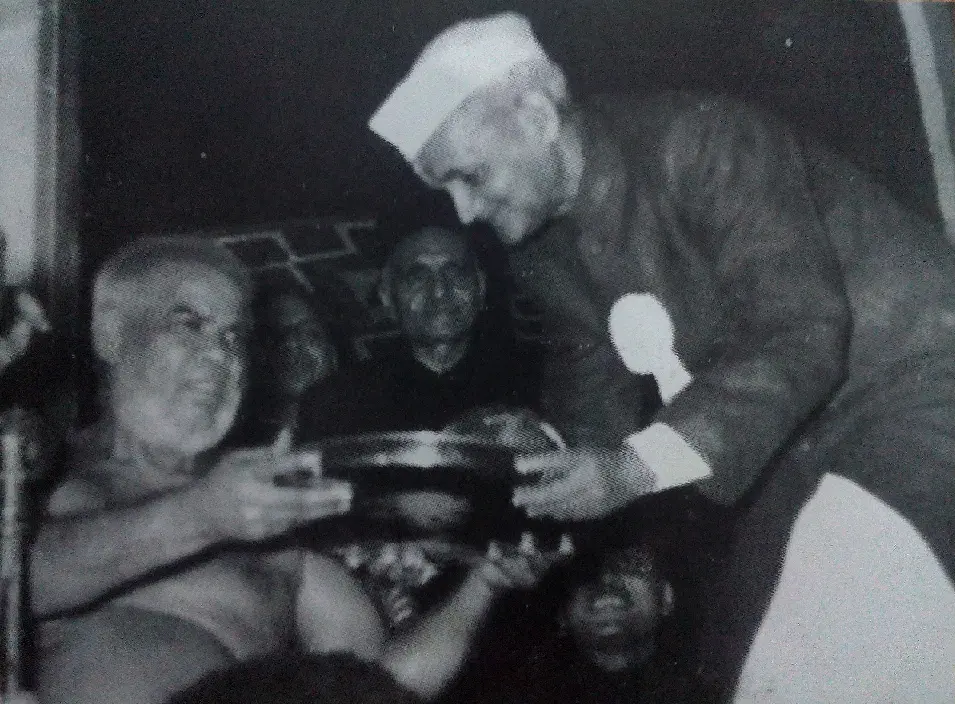an older man seated on the left receiving a round tray or plaque from another elderly man standing on the right, who is wearing a traditional Indian cap and formal attire.