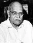 photograph of an elderly man with a bald head, glasses, and a serious expression, dressed in a light-colored shirt and seated indoors.