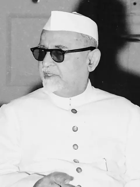 an elderly man wearing a traditional white Indian Nehru jacket and Gandhi cap, with dark sunglasses and a neatly trimmed white beard, looking slightly to the side in a formal setting.