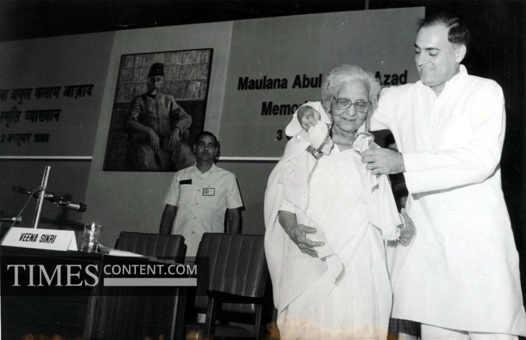 Prime Minister Rajiv Gandhi honoring Aruna Asaf Ali with a rose during the Maulana Abul Kalam Azad Memorial Lecture, as she smiles and stands on stage in a white sari.