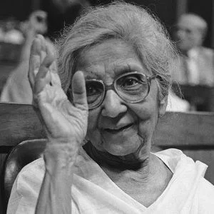Elderly Aruna Asaf Ali wearing glasses and a white sari, smiling warmly and raising her hand in greeting.