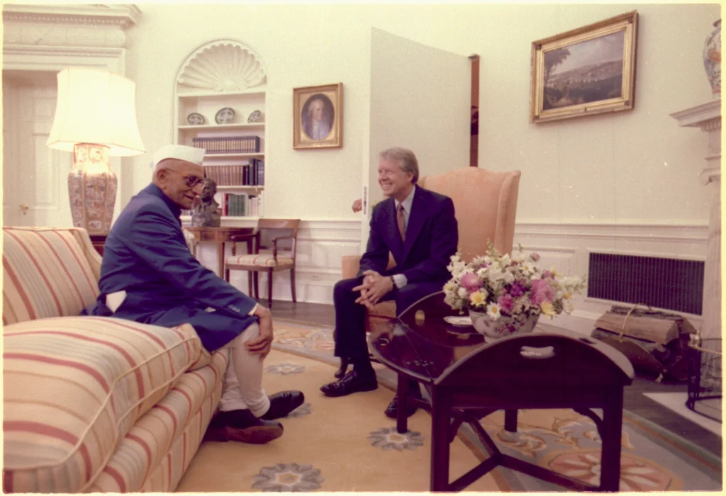 An Indian leader wearing a white Gandhi cap and blue traditional attire sits on a striped sofa, smiling and conversing with U.S. President Jimmy Carter, who is seated across from him in a formal suit, inside a well-decorated office, likely the White House.
