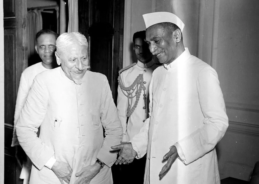 two Indian men in traditional attire engaged in a friendly conversation, with two other men in the background. The man on the left has white hair and a mustache, and the man on the right is wearing a Gandhi cap and smiling warmly.