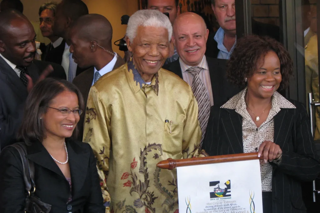 Nelson Mandela, wearing a gold patterned shirt, smiling and standing between two women, one on each side, who are also smiling. The woman on the right is holding a scroll with the logo and text of the City of Tshwane.