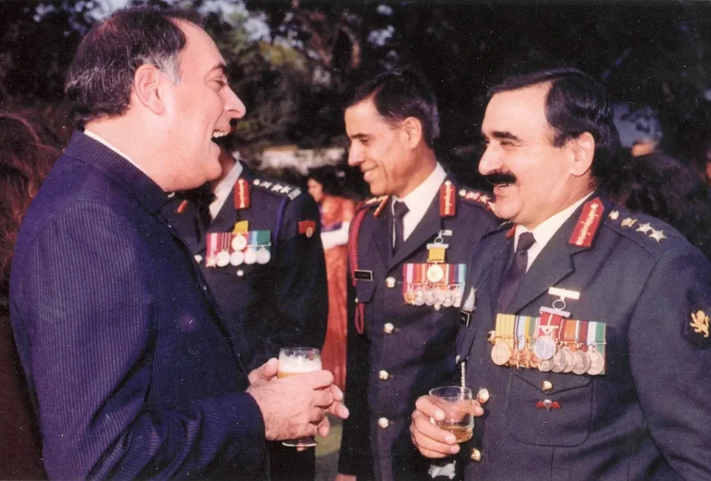 a man in a dark suit holding a drink is laughing. Two men in decorated military uniforms stand on the right, also holding drinks and smiling.