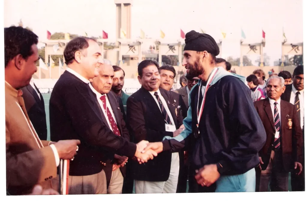 Rajiv Gandhi, former Prime Minister of India, smiling and shaking hands with an athlete wearing a medal and a turban at an outdoor event, surrounded by several men in suits.
