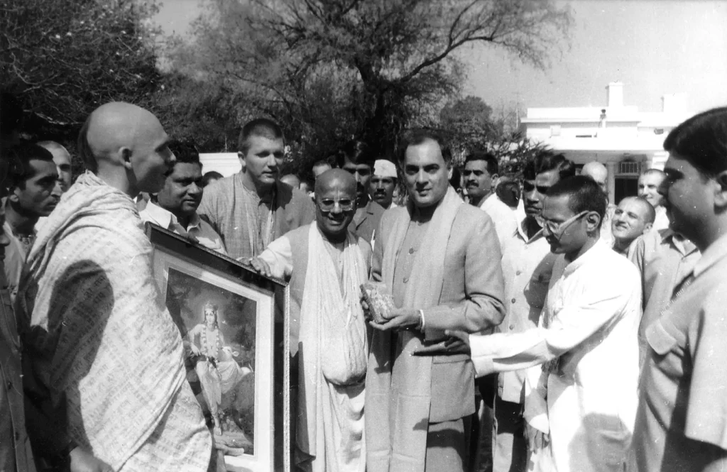 Rajiv Gandhi, former Prime Minister of India, standing outdoors surrounded by a group of men. One man is holding a framed religious painting, while Rajiv Gandhi holds a small object, possibly a gift.