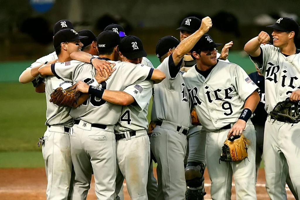 A group of Rice University baseball players hugging and celebrating a win on the field in their light gray uniforms.