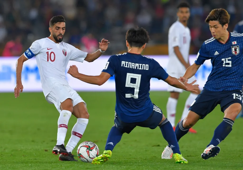 Football players from Qatar and Japan battling for the ball during an international match, with Qatar's captain Hassan Al-Haydos in possession.