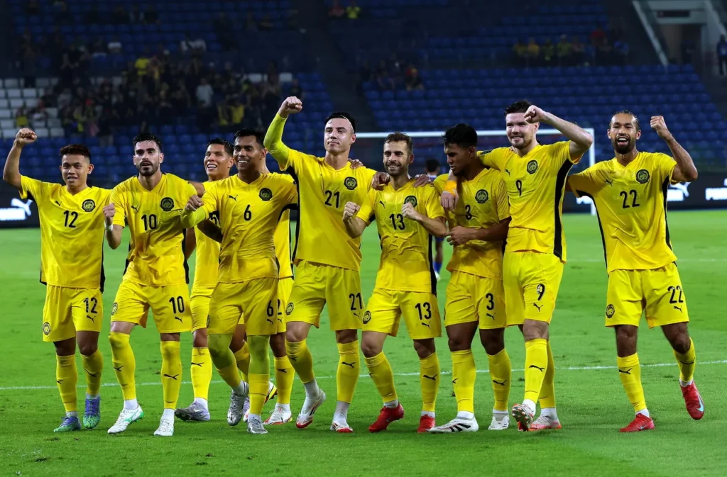 A group of Malaysian national football team players in yellow jerseys celebrating a goal or victory on the pitch.