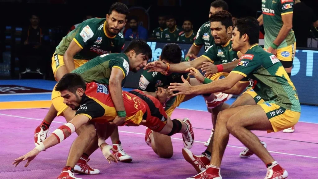 A fierce moment in a professional Kabaddi match with one raider, wearing a red and black jersey, being tackled by multiple defenders wearing green and yellow jerseys in a tight struggle.