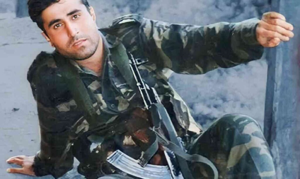 The image shows a soldier wearing a camouflage uniform, sitting on the ground with a rifle secured across his chest. His expression is focused and determined, and the background suggests a rugged or combat-like environment.
