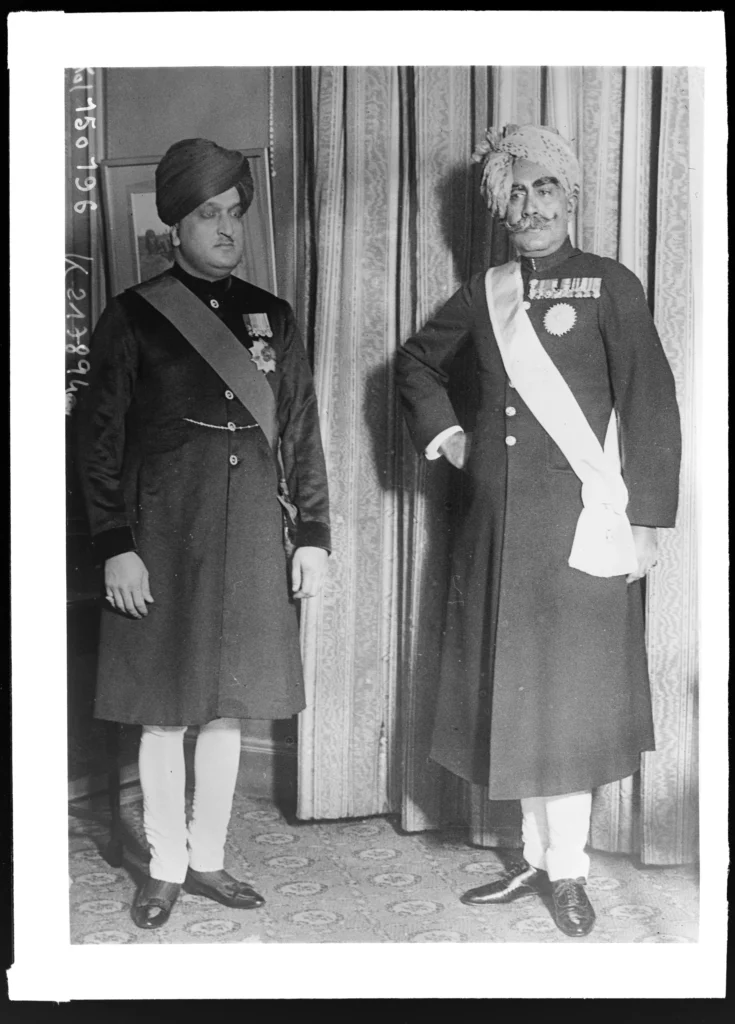 Historic black-and-white photograph of two Indian princely rulers wearing ceremonial attire and medals, standing indoors during the colonial era